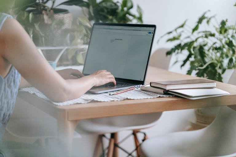 A woman typing on a laptop surrounded by plants and notebooks at her home workspace, creating a tranquil and focused environment.