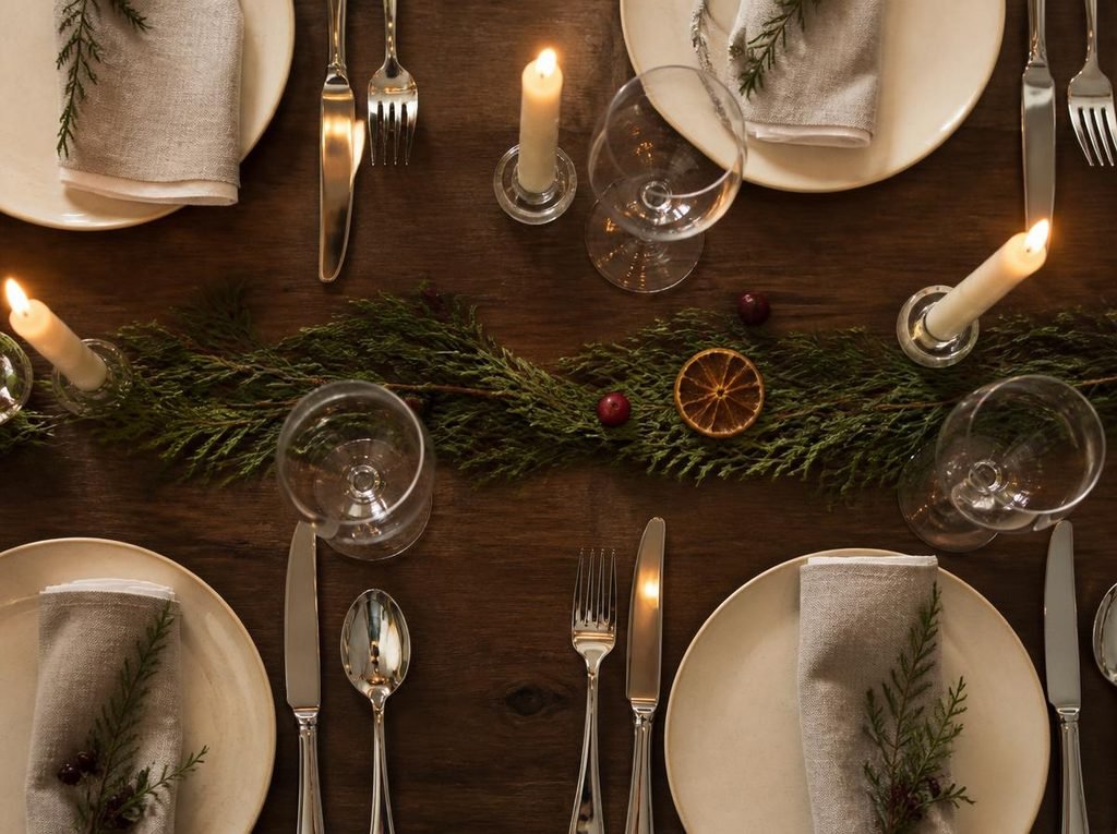 Polished stainless steel flatware reflecting warm candlelight beside cream dinnerware on an elegant Christmas table.
