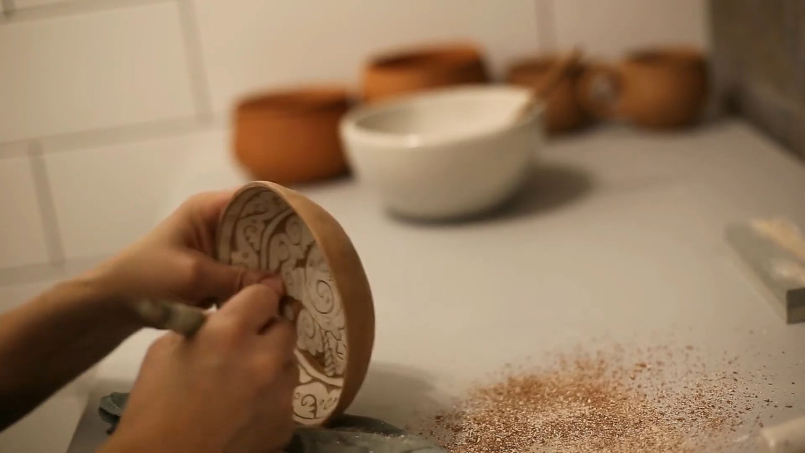Hands carving areas of pale coating from a small plate to expose the brown clay beneath.