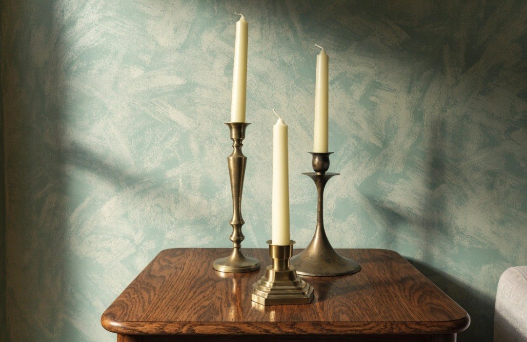 Three mismatched vintage metal candlesticks arranged at different heights with matching ivory LED taper candles on a wooden sideboard.
