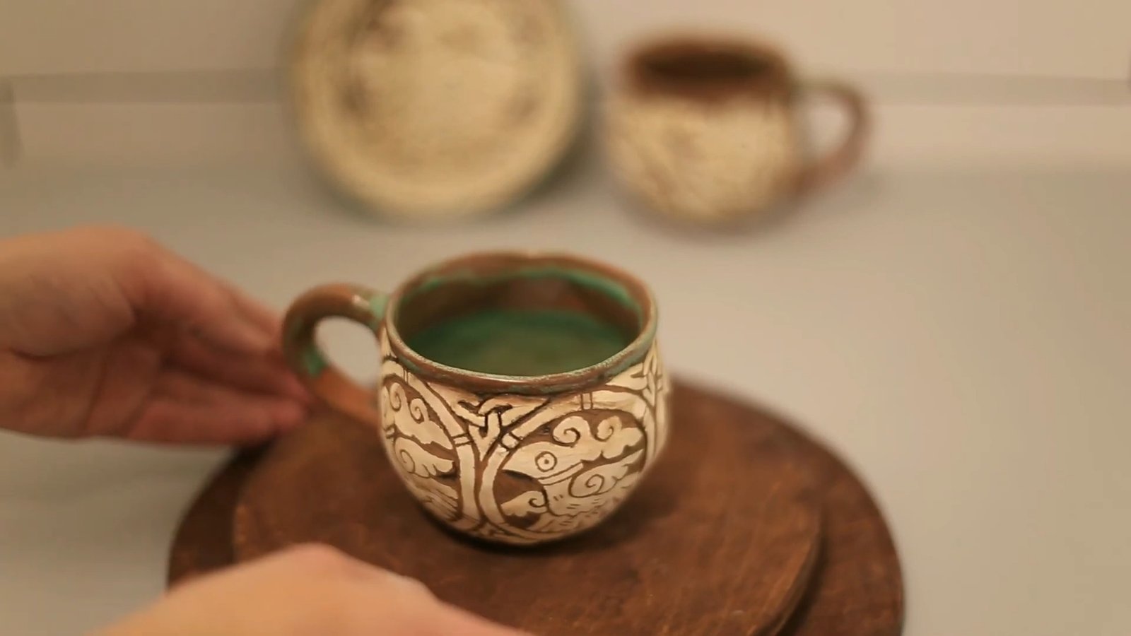 Handmade sgraffito mug with cream and brown curving motifs and a glossy green interior, shown on a wooden turntable.
