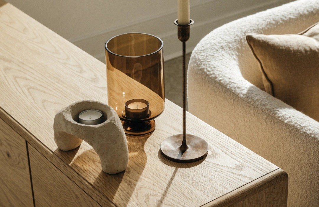 Beige ceramic LED candleholder, tan glass lantern and brown metal candlestick styled together on an oak sideboard in a neutral living room.