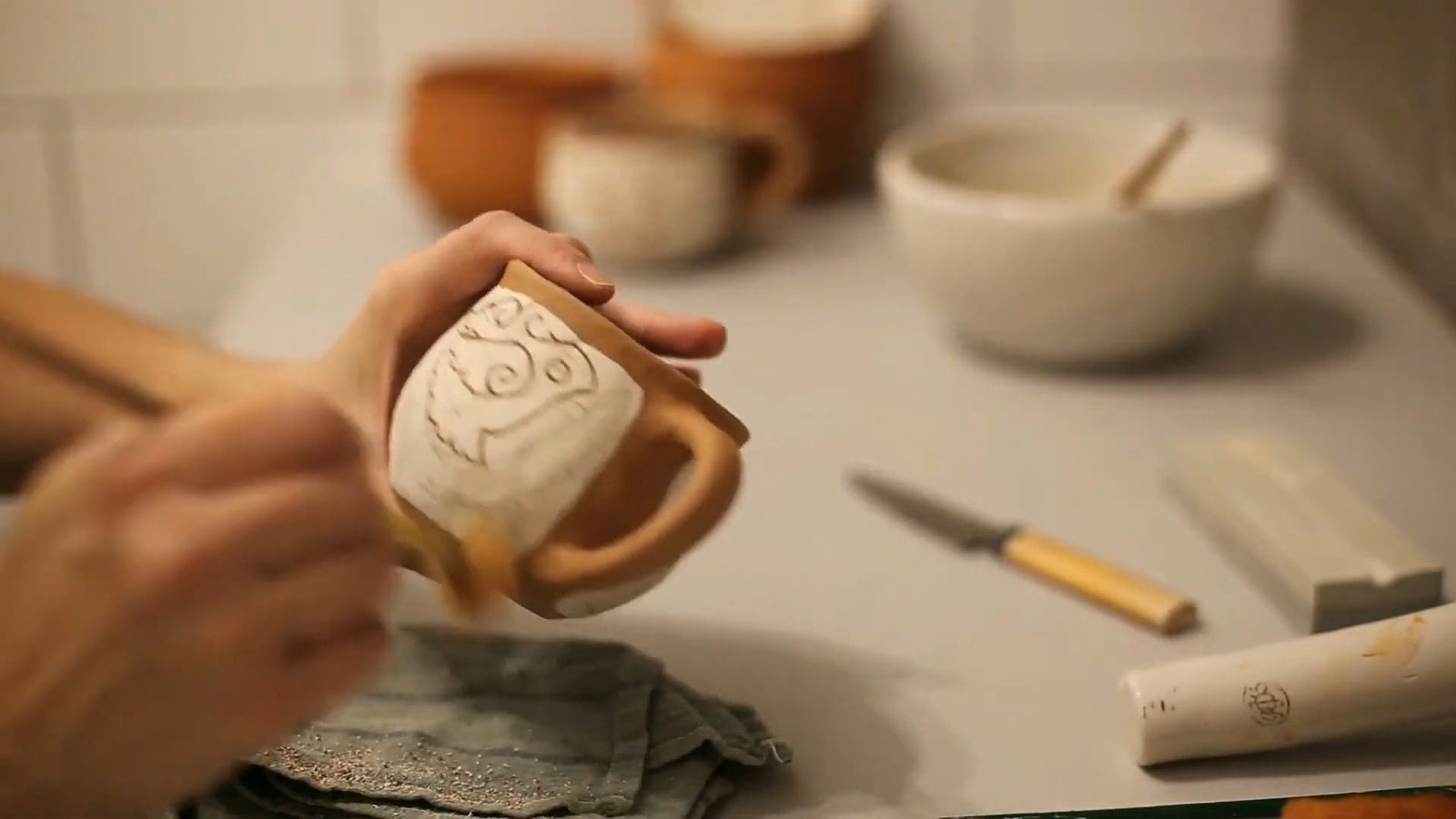 Hands working on a curving outline across the pale coating of a handmade brown clay mug.